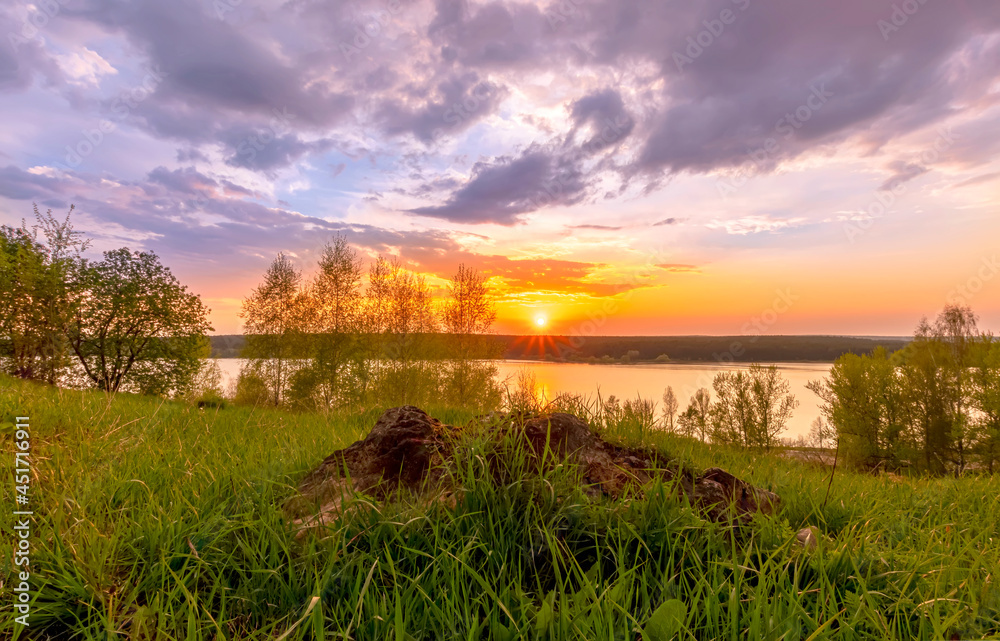 Scenic view at beautiful sunset on a shiny lake with old rough stone on the foreground, green grass, birch trees, golden sun rays, calm water ,nice cloudy sky on a background, spring landscape