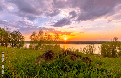 Scenic view at beautiful sunset on a shiny lake with old rough stone on the foreground, green grass, birch trees, golden sun rays, calm water ,nice cloudy sky on a background, spring landscape