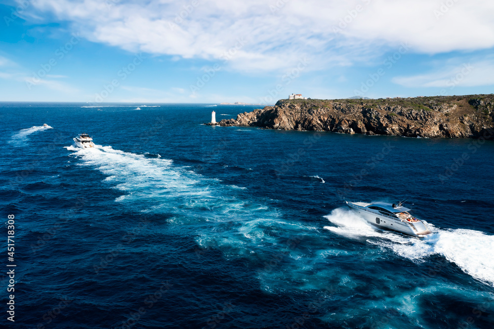 View from above, stunning aerial view of some yachts sailing on a blue sea in the foreground and a rocky coastline with a lighthouse in the distance. Cala del faro, Sardinia, Italy. View from above, stunning aerial view of some yachts sailing on a blue sea in the foreground and a rocky coastline with a lighthouse in the distance. Cala del faro, Sardinia, Italy.