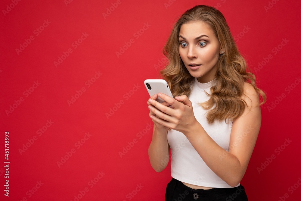 Amazed shocked beautiful young blonde woman wearing white t-shirt isolated over red background using smartphone and chatting via mobile phone looking at gadjet screen