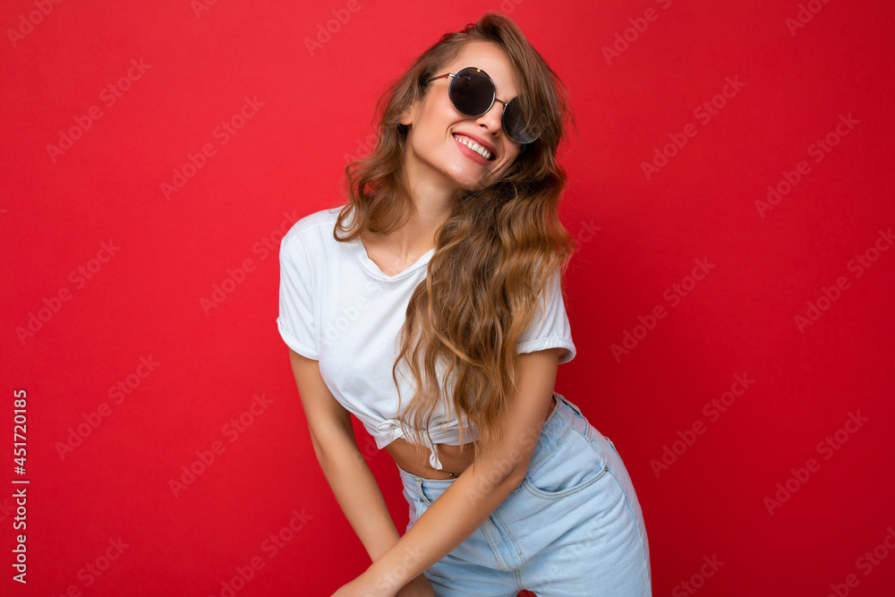 Photo shot of beautiful happy smiling young blonde woman wearing casual clothes and stylish sunglasses isolated over colorful background looking at camera Photo shot of beautiful happy smiling young blonde woman wearing casual clothes and stylish sunglasses isolated over colorful background looking at camera