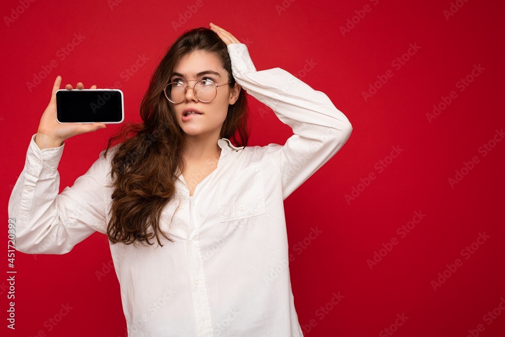 Naklejka premium photo of sexy beautiful young brunette woman wearing white shirt and optical glasses isolated over red background holding in hand and showing mobile phone with empty screen for cutout looking at