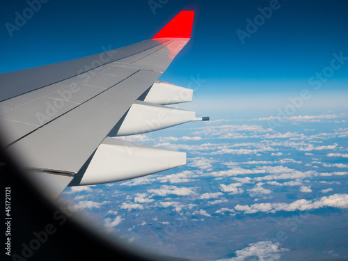 View from passenger cabin window on wing of flying plane against background of blue sky and white clouds