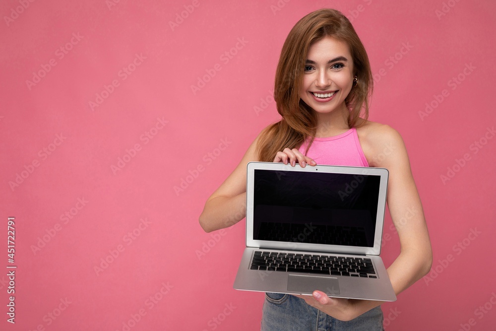 Naklejka premium Beautiful young blond woman holding computer laptop with empty monitor display wearing pink crop top looking at camera isolated on pink background