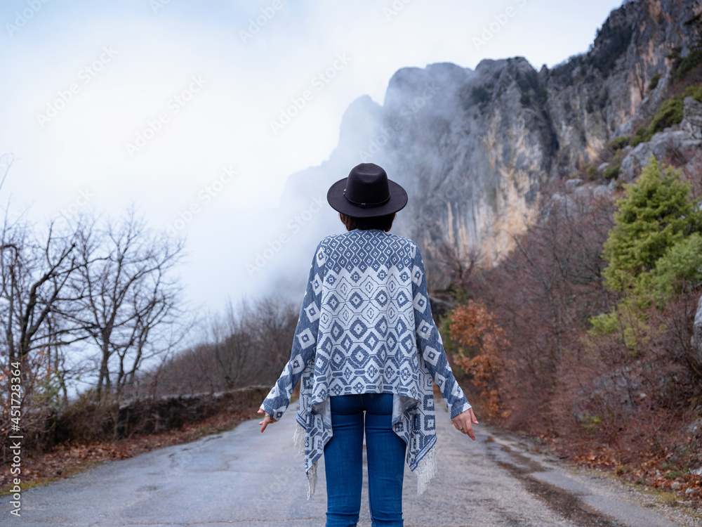 Back view of Stylish hipster woman in hat and poncho walking down a mountain road. The concept of travel and wanderlust. An amazing atmospheric moment Back view of Stylish hipster woman in hat and poncho walking down a mountain road. The concept of travel and wanderlust. An amazing atmospheric moment