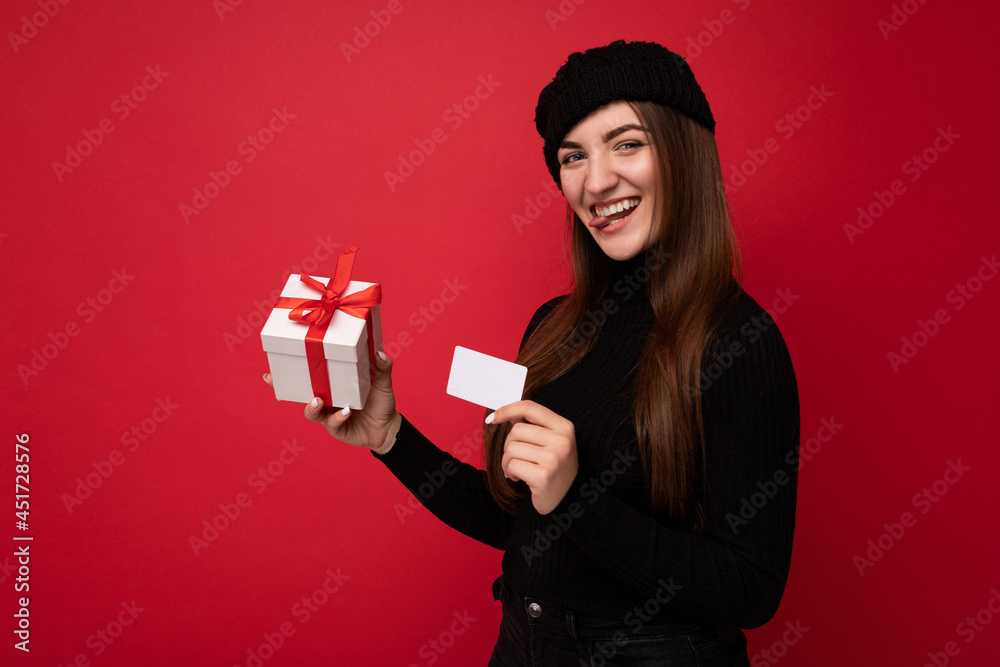 Fototapeta premium Beautiful happy young brunette woman wearing black sweater and hat isolated on red background holding credit card and gift box looking at camera