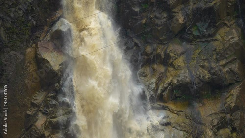 Dirty water crashing down the waterfall in slow motion along rocky wet cliff wall - Banos,Ecuador