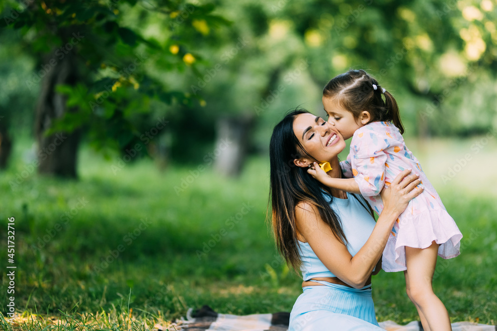 Fototapeta premium Mother and daughter lying on the lawn. Daughter kiss her mother on the cheek. Family in the city park outdoors. Happiness of motherhood and childhood