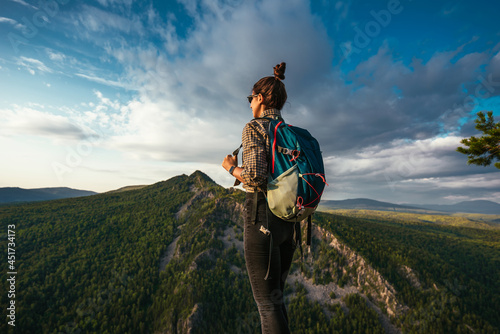 A young tourist girl with a backpack enjoys the sunset from the top of the mountain. A traveler on the background of mountains. A tourist with a backpack is standing on the top of the mountain