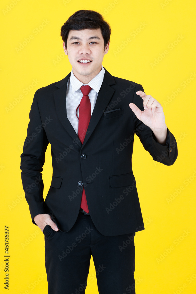 Vertical portrait shot of young Asian man in a black long sleeve suit with hand in pocket posing presenting and showing the empty space with the fingers isolated with yellow background in studio. Vertical portrait shot of young Asian man in a black long sleeve suit with hand in pocket posing presenting and showing the empty space with the fingers isolated with yellow background in studio.