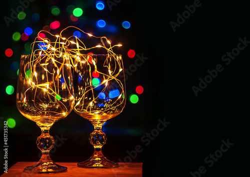 Crystal wine glasses with golden glowing garland on Christmas table bokeh light background copy space for text. Romantic New Years eve dinner champagne couple in love. Magic winter holidays atmosphere