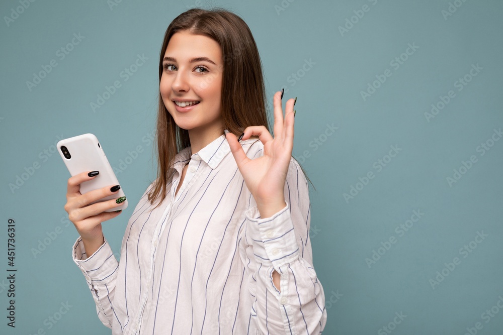 Side-profile Photo of smiling beautiful young blonde woman wearing casual white shirt isolated over blue background wall holding smartphone and texting message looking at camera and showing okay Side-profile Photo of smiling beautiful young blonde woman wearing casual white shirt isolated over blue background wall holding smartphone and texting message looking at camera and showing okay