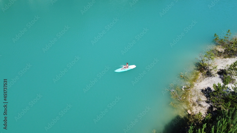 Naklejka premium A girl in a dress floats on a glanders board on a pond with bright turquoise water. Warm summer day for travel. Top view from a quadcopter. Aerial photography