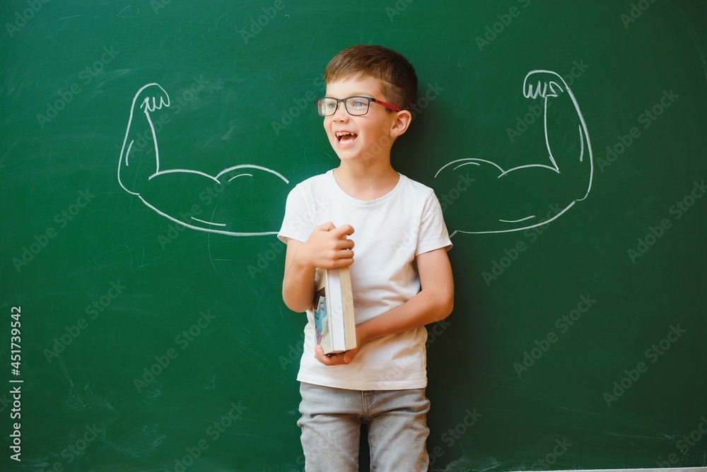 Cute child boy in school uniform and glasses. Go to school for the first time. Child with school bag and books. Kid in class room near chalkboard with muscles on it. Back to school