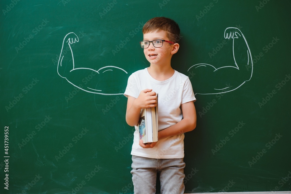 Cute child boy in school uniform and glasses. Go to school for the first time. Child with school bag and books. Kid in class room near chalkboard with muscles on it. Back to school