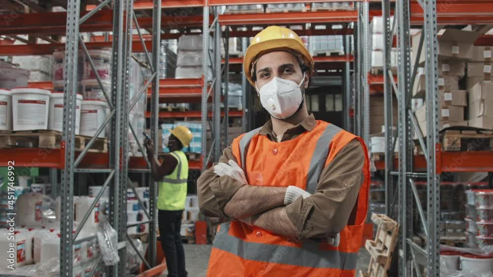 Slowmo tracking portrait of young male worker in safety vest, hard hat, gloves and face mask crossing his arms and posing for camera in warehouse of large hardware store with items stacked on shelves Slowmo tracking portrait of young male worker in safety vest, hard hat, gloves and face mask crossing his arms and posing for camera in warehouse of large hardware store with items stacked on shelves
