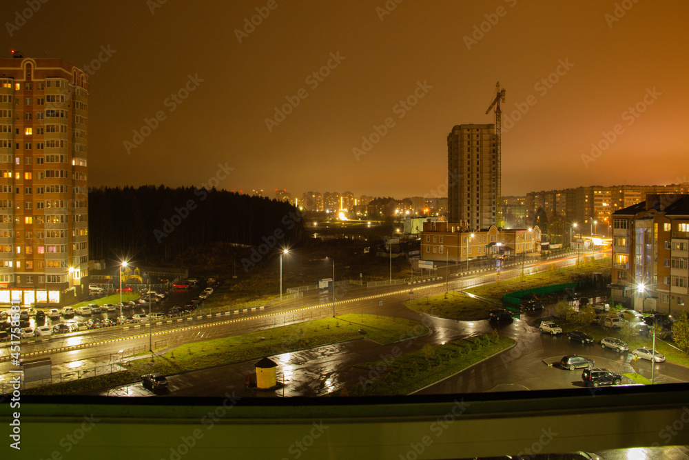 Night view of the suburb of the metropolis. Road with residential multi-storey buildings in a residential area, urban forest area, new building and parking in the foreground. Summer. From windows