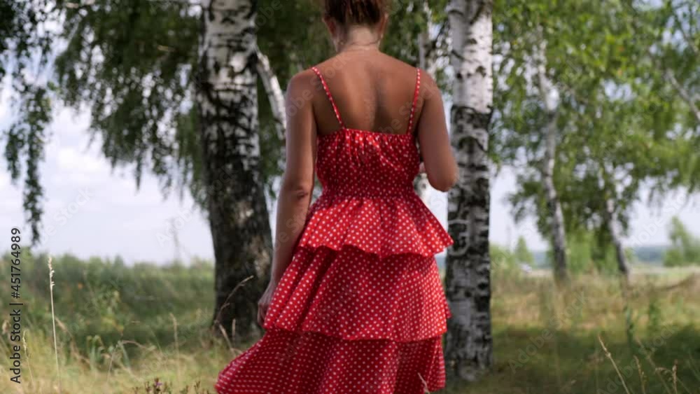 A slender girl with a topknot in a red polka-dot off-the-shoulder dress is walking through the woods among the birch trees in slow motion. This shot is from the back, medium long shot. A slender girl with a topknot in a red polka-dot off-the-shoulder dress is walking through the woods among the birch trees in slow motion. This shot is from the back, medium long shot.
