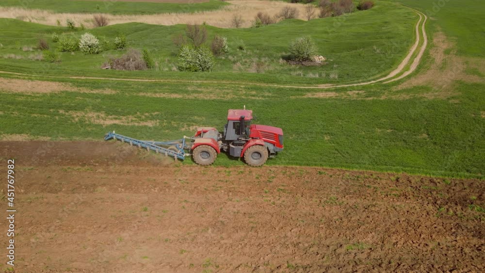 Red tractor with harrows is loosening the ground to protect soil from drying out, level its surface, destroy weeds and prepare for seasonal planting wheat, shooting with drone from height, side view. Red tractor with harrows is loosening the ground to protect soil from drying out, level its surface, destroy weeds and prepare for seasonal planting wheat, shooting with drone from height, side view.