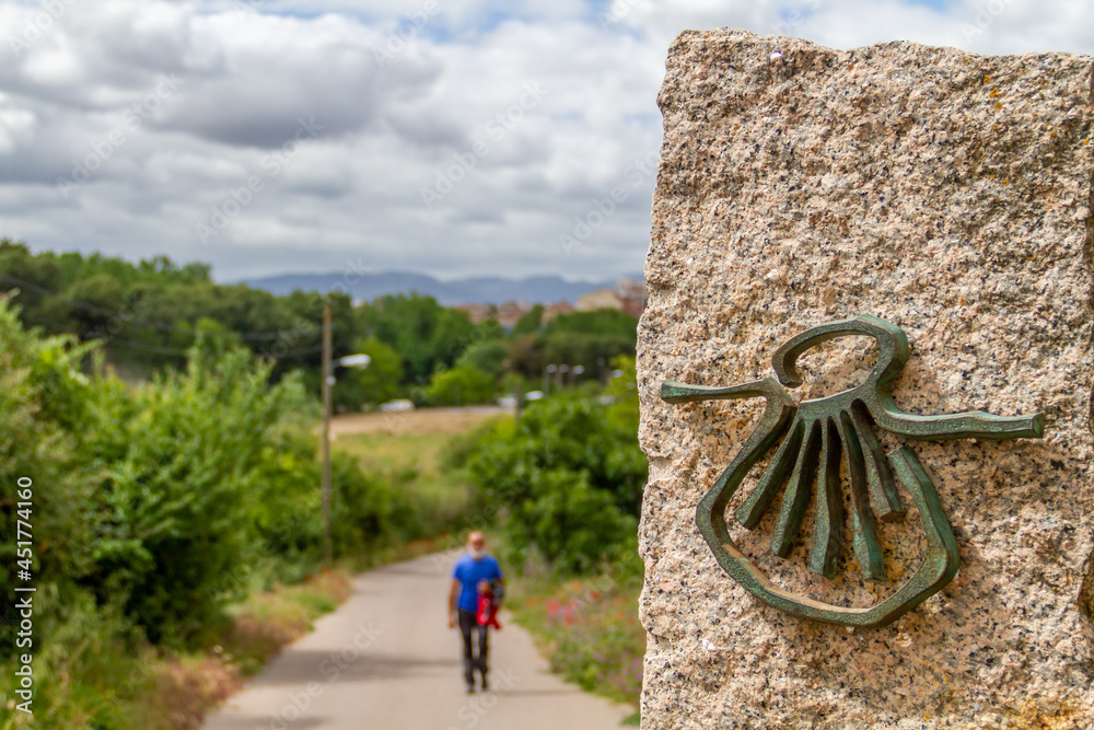 Pilgrim out of focus walks along the Camino de Santiago (Sant James Way) carrying a backpack next to a column on focus with the shell symbol of the Camino Santiago de Compostela - obrazy, fototapety, plakaty 
