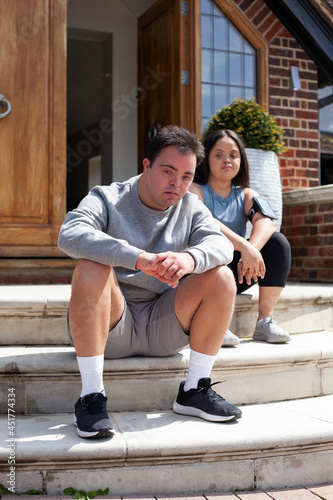 Young biracial couple with Down Syndrome in active wear and wearable tech sitting on steps looking confident