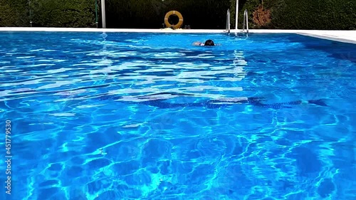 A woman in a pool swimming backward with her face looking up to the sky and moving towards the camera with simultaneous backstrokes with both arms. An orange buoyancy aid rubber ring at the back photo