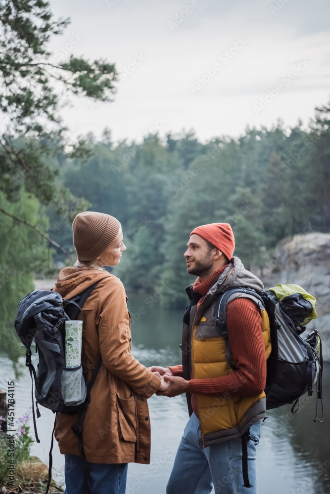 young and joyful couple holding hands while looking at each other near lake in forest young and joyful couple holding hands while looking at each other near lake in forest