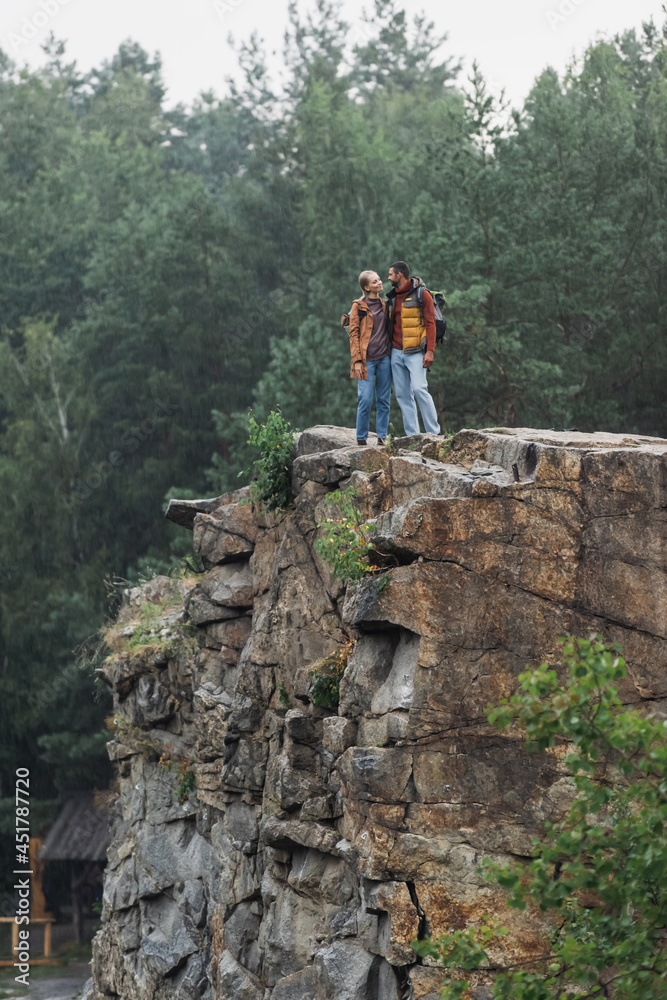 cheerful couple with hiking sticks hugging on rocky cliff