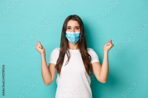 Concept of pandemic, covid-19 and social distancing. Image of confideny lucky girl, feeling enthusiastic, clench fists and rejoicing, wearing medical mask, standing over blue background photo