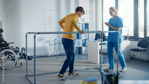 Hospital Physical Therapy Room: Strong Patient with Injury Walking Holding for Parallel Bars, Physiotherapist Encourages Assists, Helps Disabled Person Do Rehabilitative Physiotherapy.