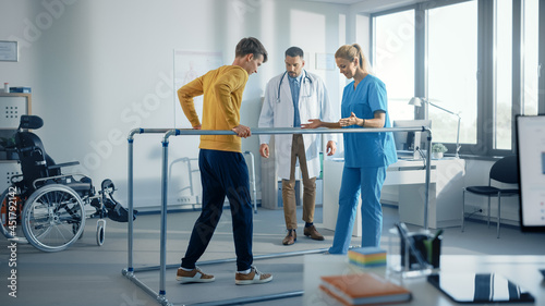 Fototapeta Naklejka Na Ścianę i Meble -  Hospital Physical Therapy: Strong Patient with Injury Making First Steps, Walks Holding for Parallel Bars. Physiotherapist, Rehabilitation Doctor Assist, Help Disabled Person.