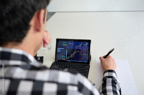 stock market data on a screen and young man takes notes on a blank paper