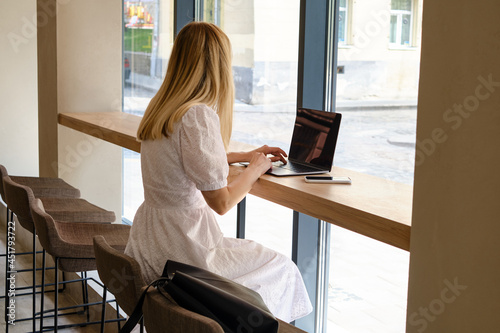 The girl sits at the table and works at a laptop and a smartphone. Remote work from home, cafe, business space. Business lady at work. Business background. Home office © Iuliia Pilipeichenko