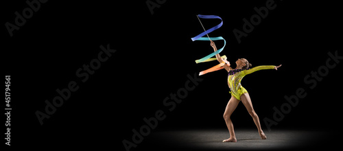 Flyer with little girl, rhythmic gymnastics artist in bright stage costume isolated on dark studio background in ray of light. Concept of sport, action, aspiration, active lifestyle