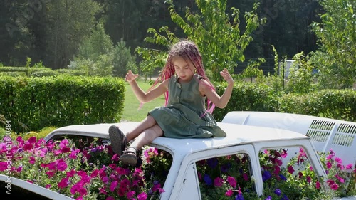 A girl with pink zizi afro - pigtails sits on the roof of a decorative car with flowers growing from inside and plays with her hair, scattering collected pigtails into a bun. Landscape Design Ideas photo