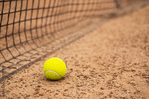 A new tennis ball is lying on a sand court near the net.