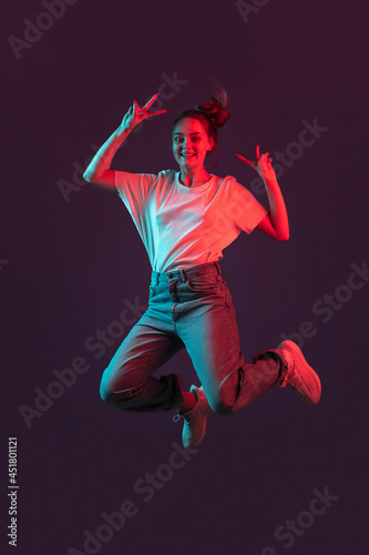 Vertical portrait of beautiful girl in casual clothes jumping isolated on dark purple studio background in red neon light, filter. Concept of human emotions, youth culture