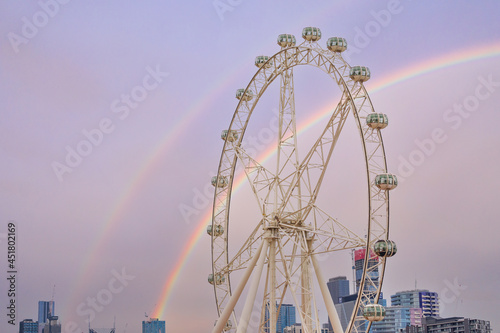 Horizontal shot of a double rainbow behind a large modern ferris wheel and city skyline