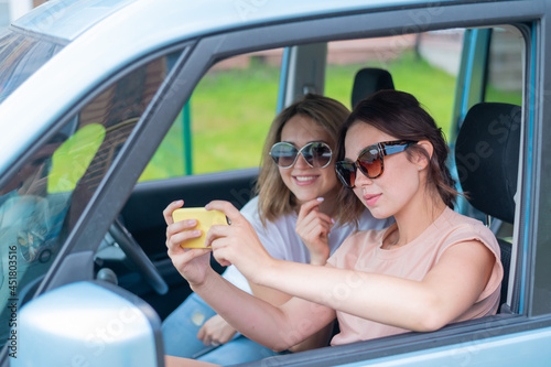 Two happy girlfriends go on a trip. Women are driving in a car and taking a selfie on a mobile phone