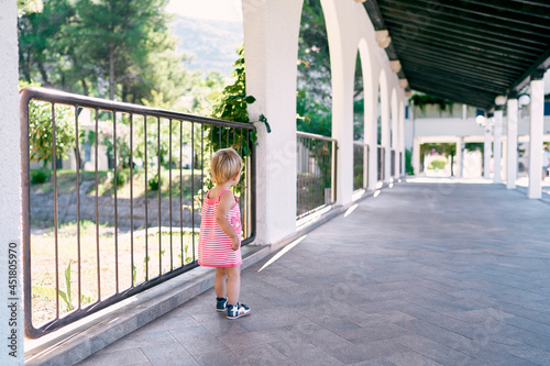 Little girl stands near a fence in a long pavilion in the park. Back view