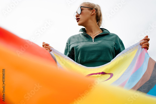 Fashionable young woman with glasses and red lipstick holds rainbow flag, international symbol of LGBTQ+ community, gay, lesbian, people of color and transperson. Equal rights and freedoms for LGBT photo