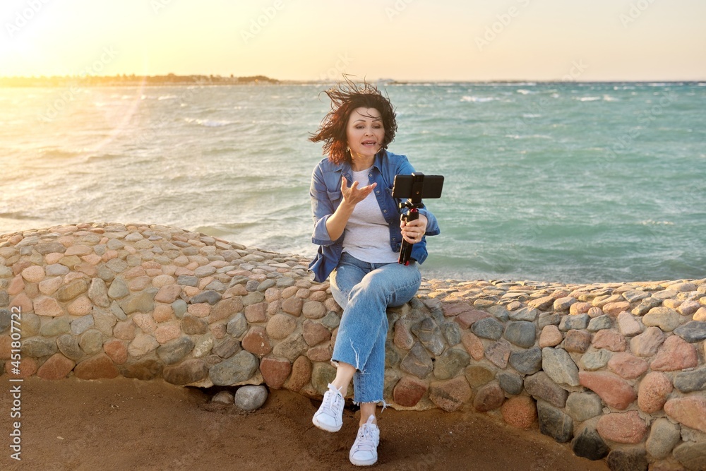 Middle-aged female making online video call talking laughing, sunset seascape background