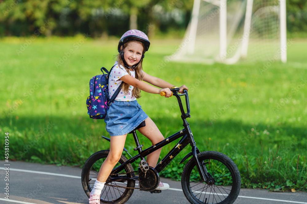 little schoolgirl girl with backpack wears pink protective helmet in park on sunny summer day. child rides bicycle to school. Safe riding is fun childhood. An active game. first day of school. little schoolgirl girl with backpack wears pink protective helmet in park on sunny summer day. child rides bicycle to school. Safe riding is fun childhood. An active game. first day of school.