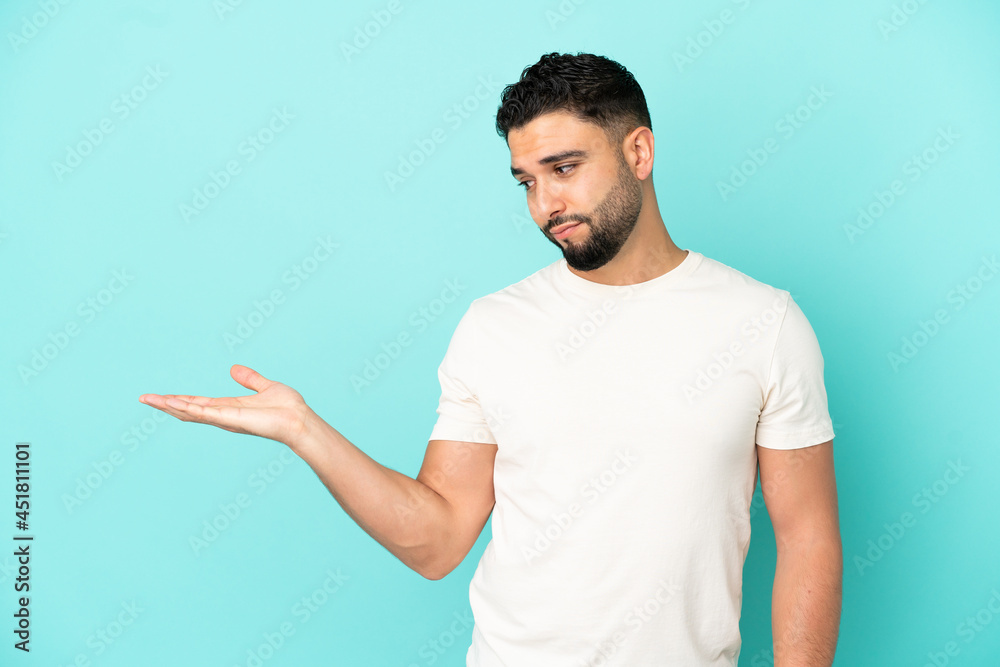 Young arab man isolated on blue background holding copyspace with doubts Young arab man isolated on blue background holding copyspace with doubts