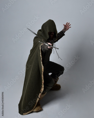 Full length portrait of young handsome man wearing medieval Celtic adventurer costume with hooded cloak, holding sword, isolated on studio background.