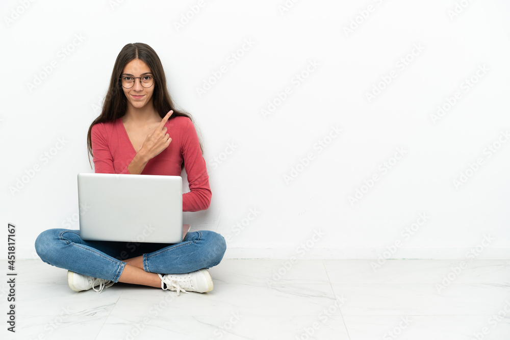 Young French girl sitting on the floor with her laptop pointing to the side to present a product