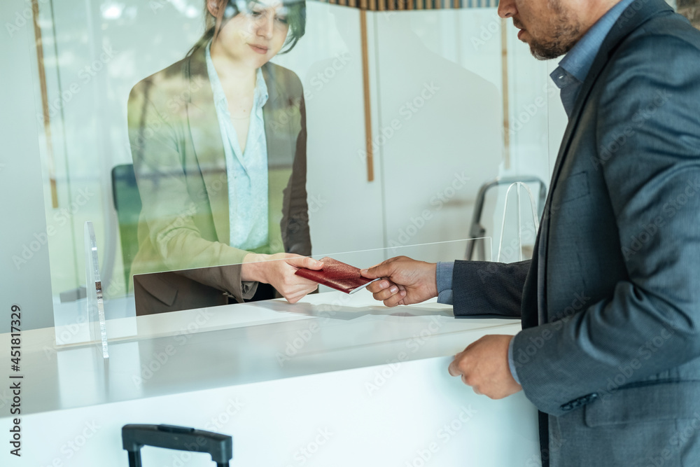 Obraz premium Unrecognizable Businessman Handing Over Passport to Hotel Receptionist. An anonymous business man passing documents to woman concierge on hotel reception with sneeze guard protection. 