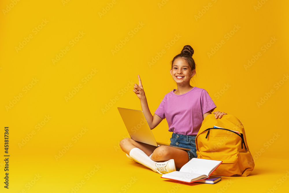 happy schoolgirl with backpack, laptop and books is sitting on yellow background
