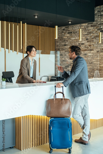 Female Hotel Receptionist Assisting Businessman for Checking In.
Business man with luggage talking with concierge on hotel reception with sneeze guard protection.