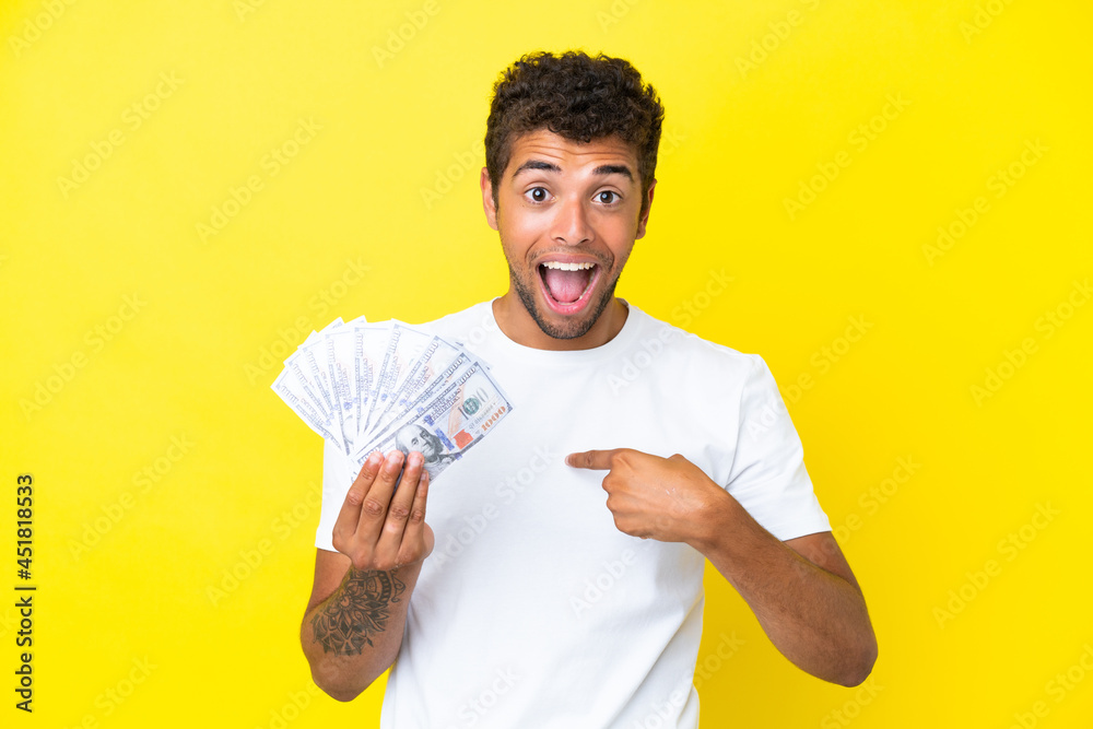 Young brazilian man taking a lot of money isolated on yellow background with surprise facial expression Young brazilian man taking a lot of money isolated on yellow background with surprise facial expression
